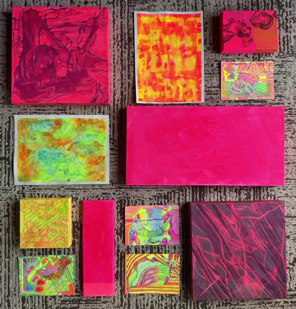 Canvases and paper pieces in progress laid out on a carpet, washed in neon pink, orange, yellow and green